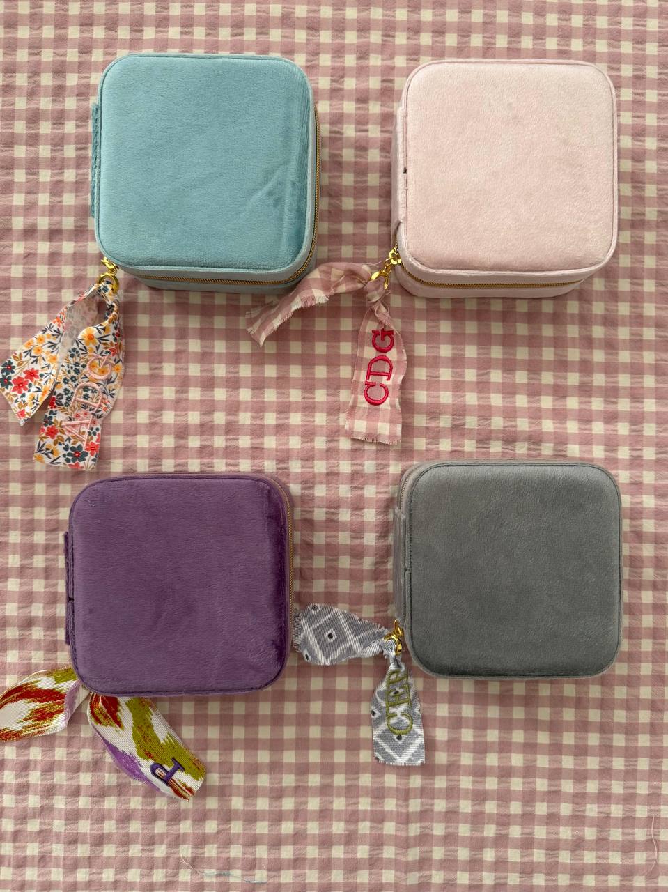 Cuatro joyeros de viaje de terciopelo en menta, rosa, morado y gris con cintas personalizadas sobre tela de cuadros vichy rosa