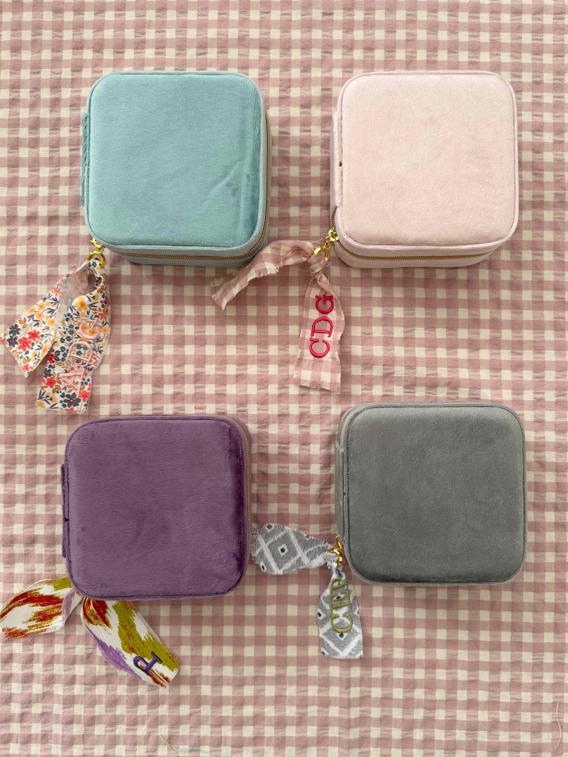 Cuatro joyeros de viaje en terciopelo desde arriba: menta, rosa, morado y gris, cada uno con su cinta personalizada